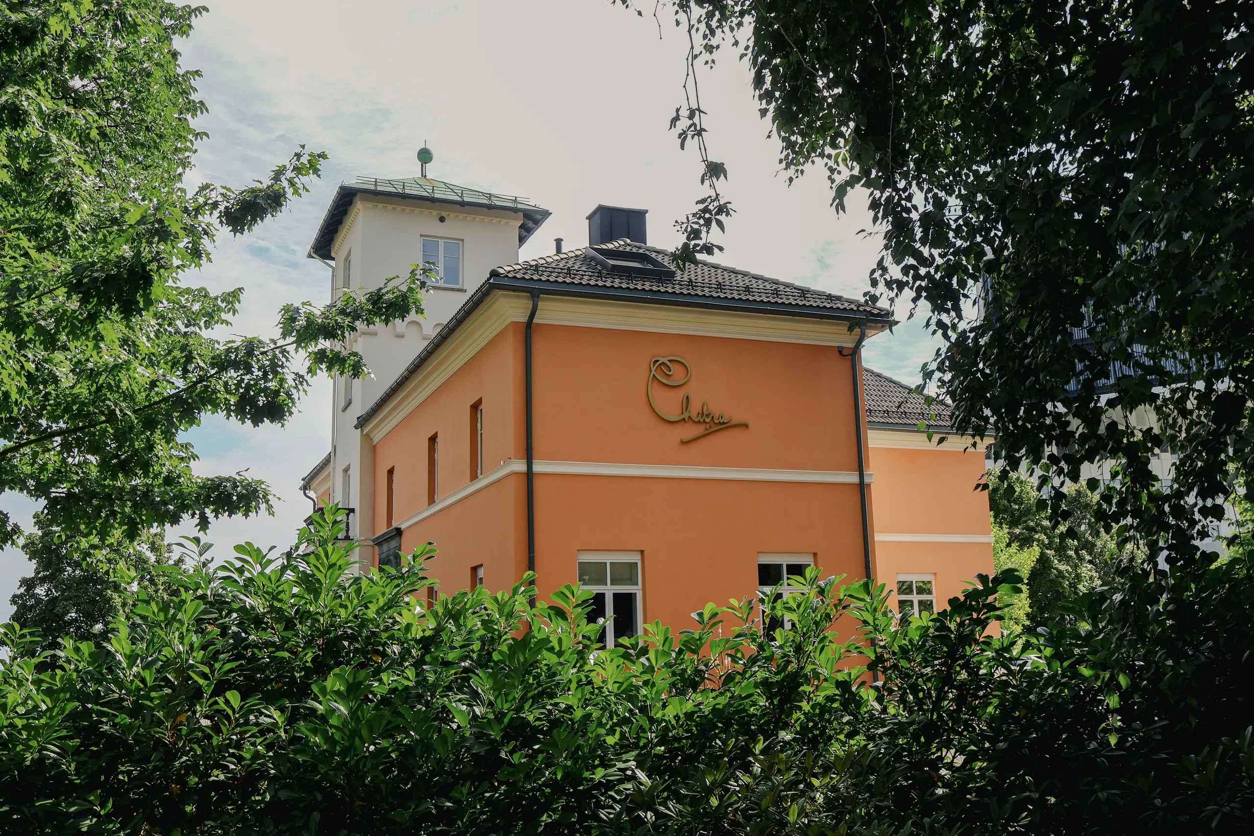 Fasaden på Chitra house med tårn og forgylt Chitra-logo, omkranset av grønne trær